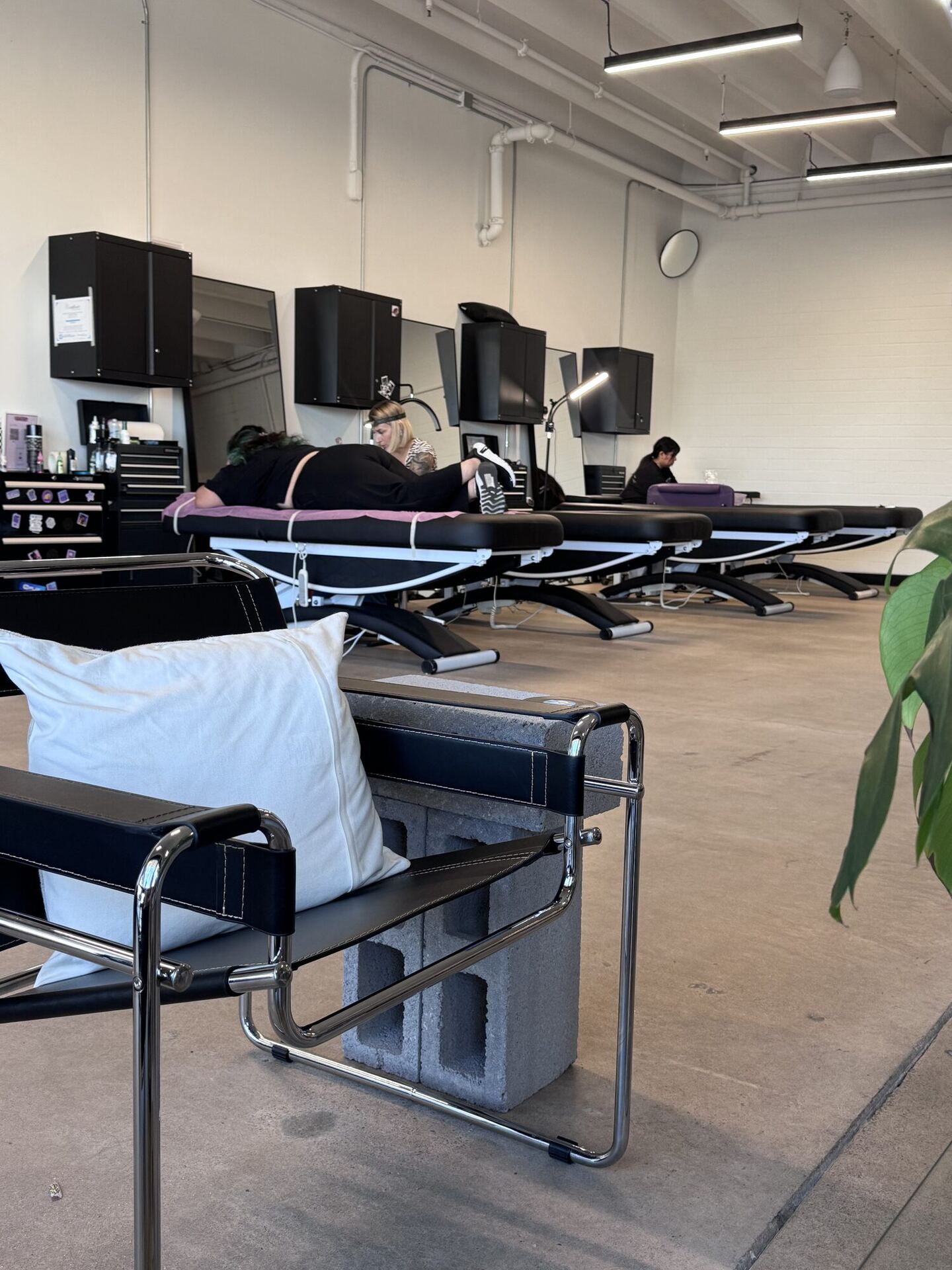 The tattoo studio at the back of the shop: a row of black tattoo beds under mirrors and cabinets, an artist bent over a client lying face down, and in the foreground a chrome-and-leather chair with a white pillow, propped on a cinder block.