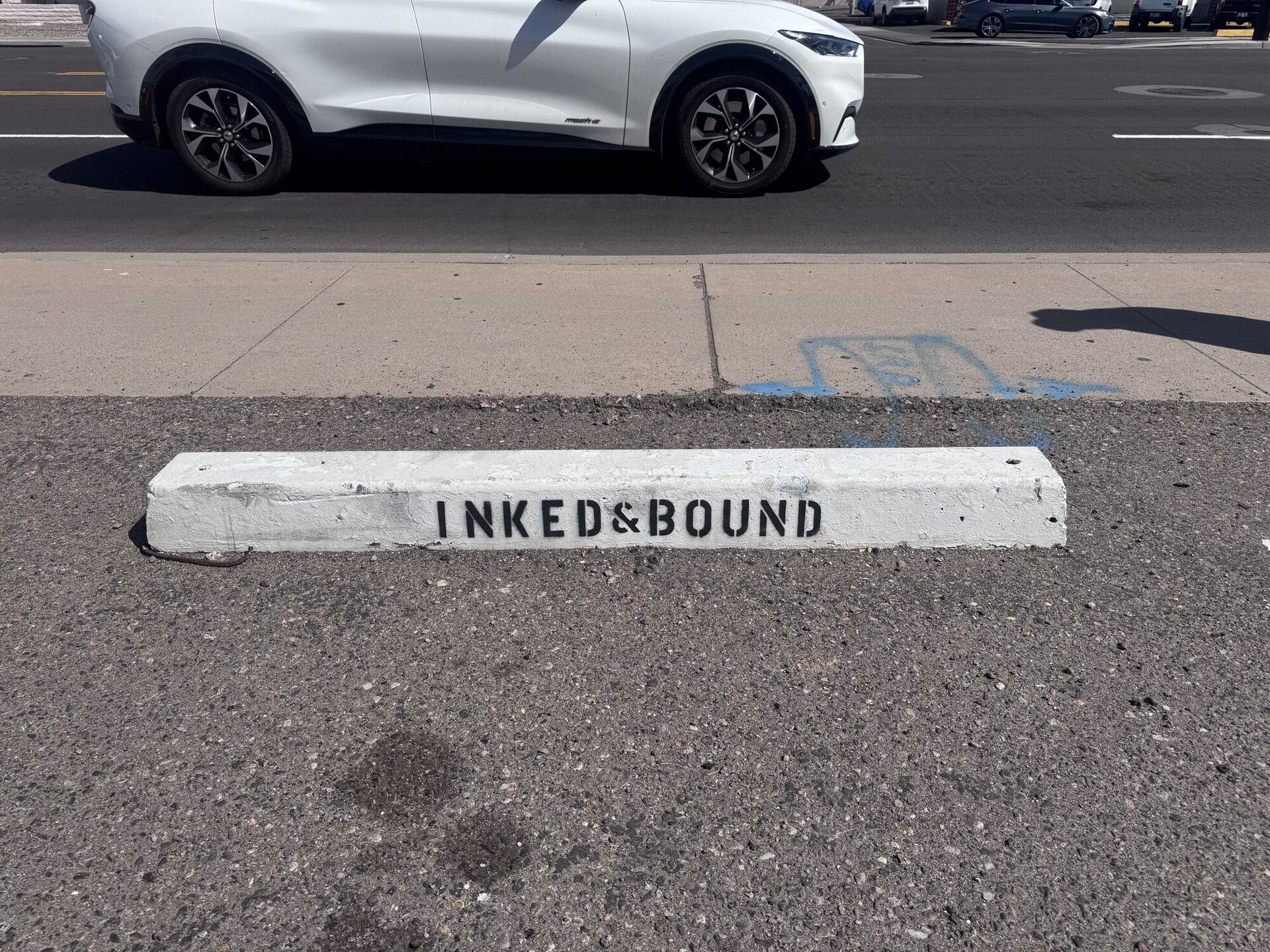 A white-painted concrete parking bumper on the asphalt, stencilled INKED&BOUND in black capitals, with a white SUV passing on the street beyond.