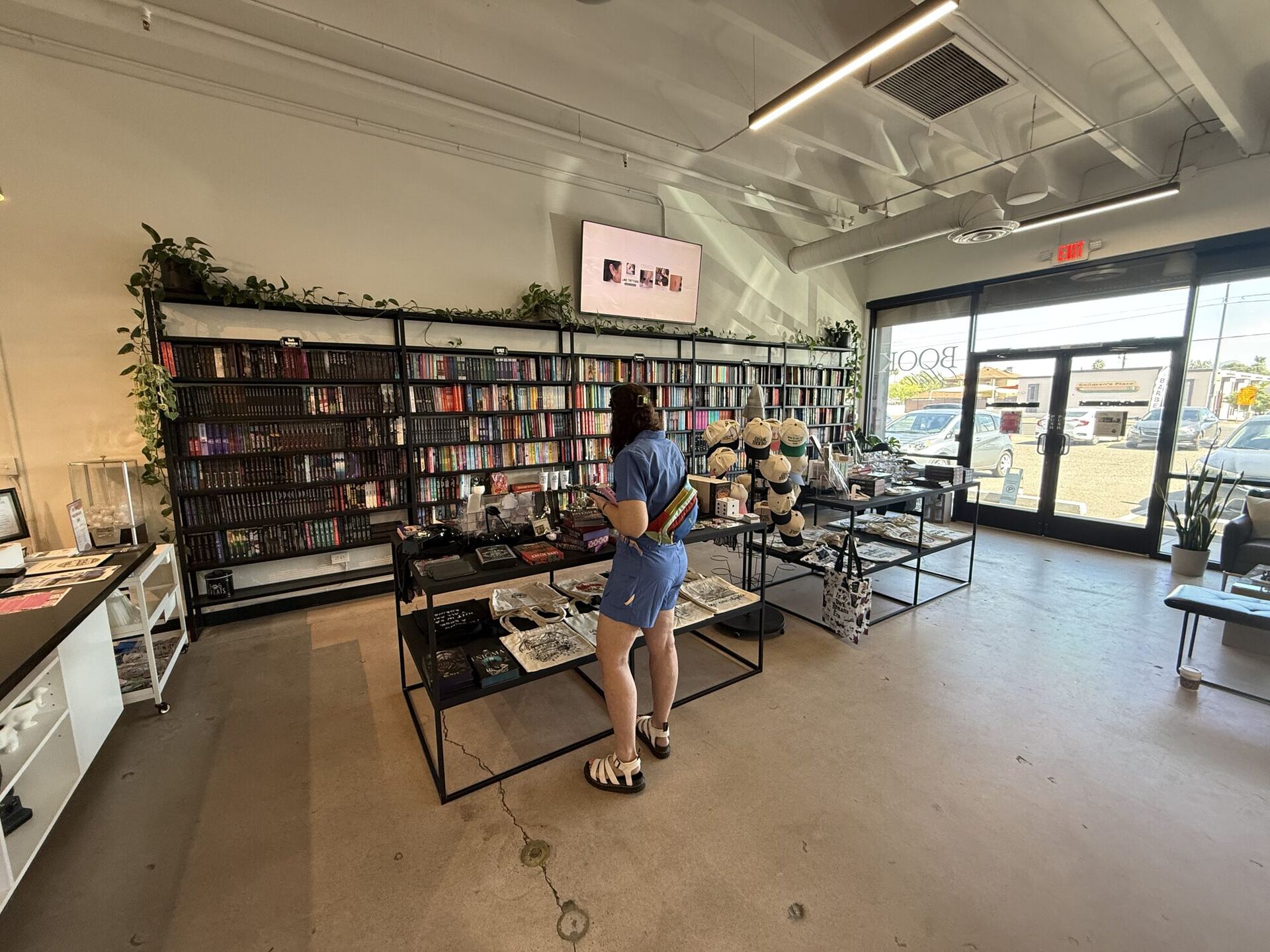 A bright, high-ceilinged shop floor. Black wire shelving runs the length of a white wall, packed with paperbacks and trailing pothos. A customer in a blue romper stands at a low merch table beside a rack of embroidered caps, and the glass storefront opens onto a sunlit car park.
