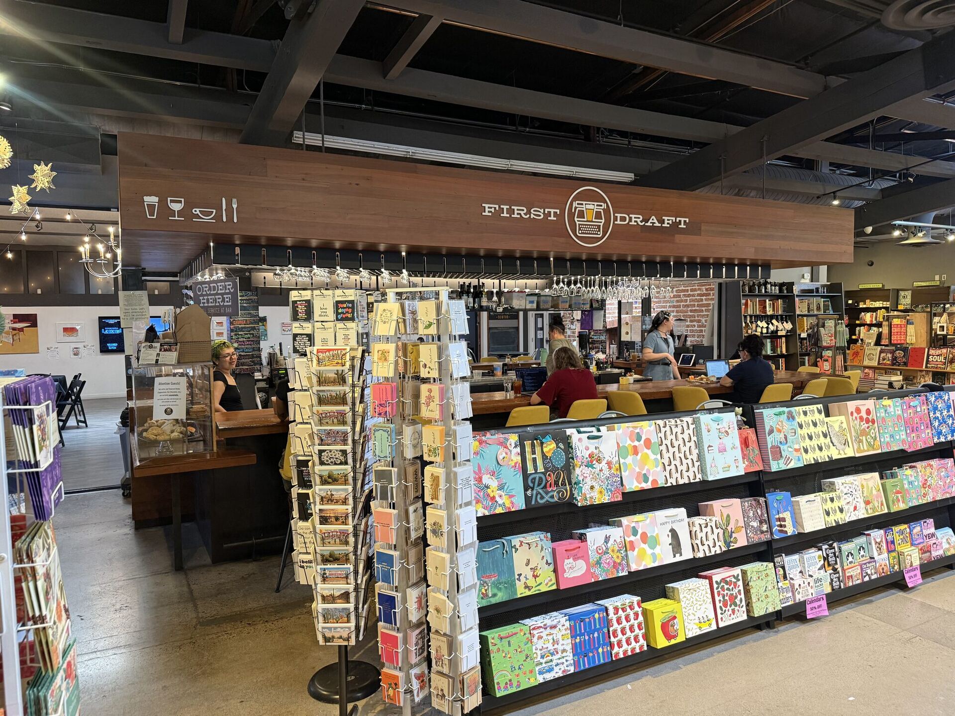 A wide view of the cafe counter under the FIRST DRAFT sign, with spinning racks of greeting cards and a wall of illustrated gift bags in the foreground and customers at the bar beyond.