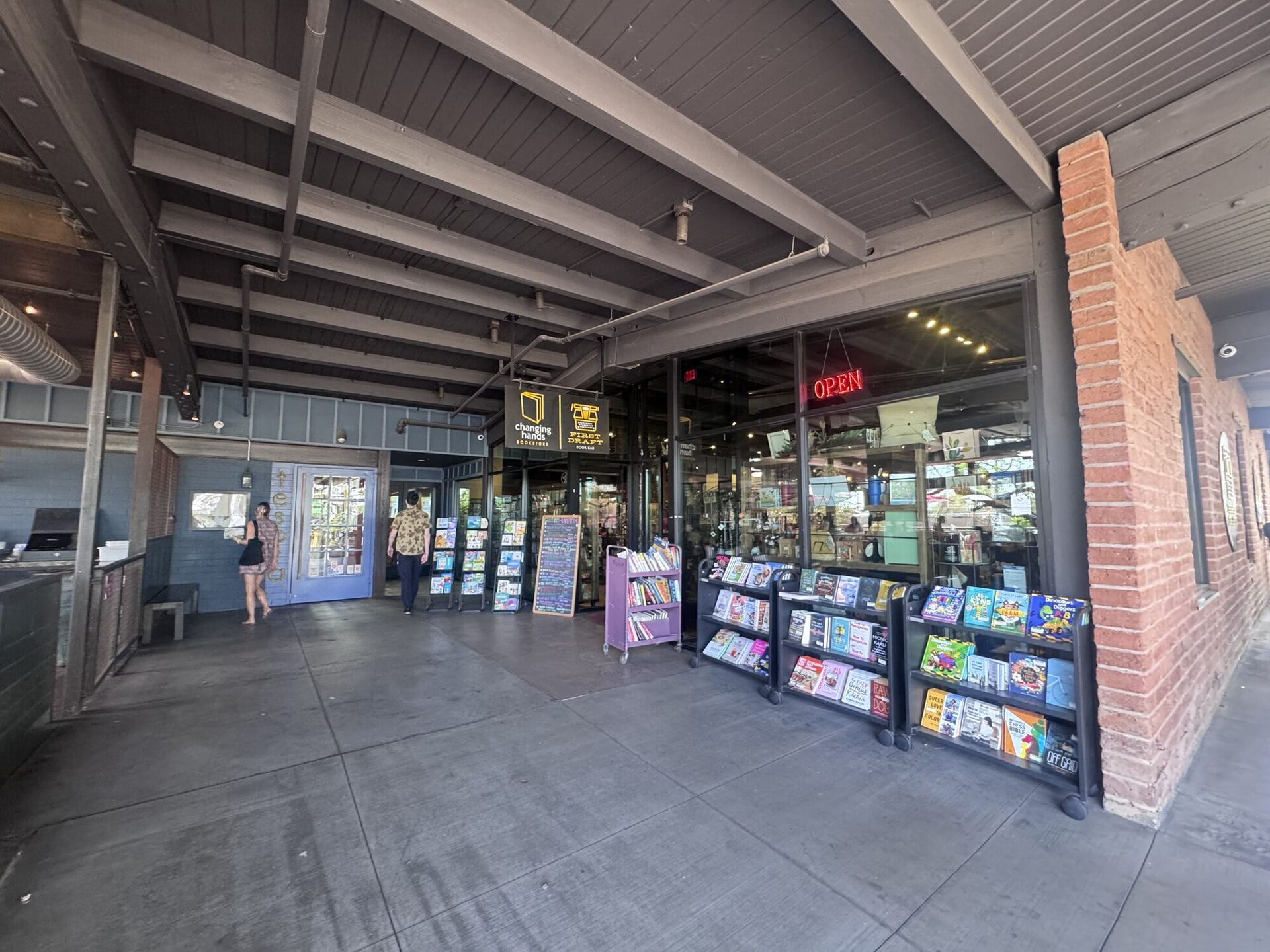 A deep covered walkway with heavy timber beams and a brick pillar. A red neon OPEN sign glows in the window, hanging signs read changing hands bookstore and FIRST DRAFT book bar, and wheeled carts of discounted books line the pavement outside.