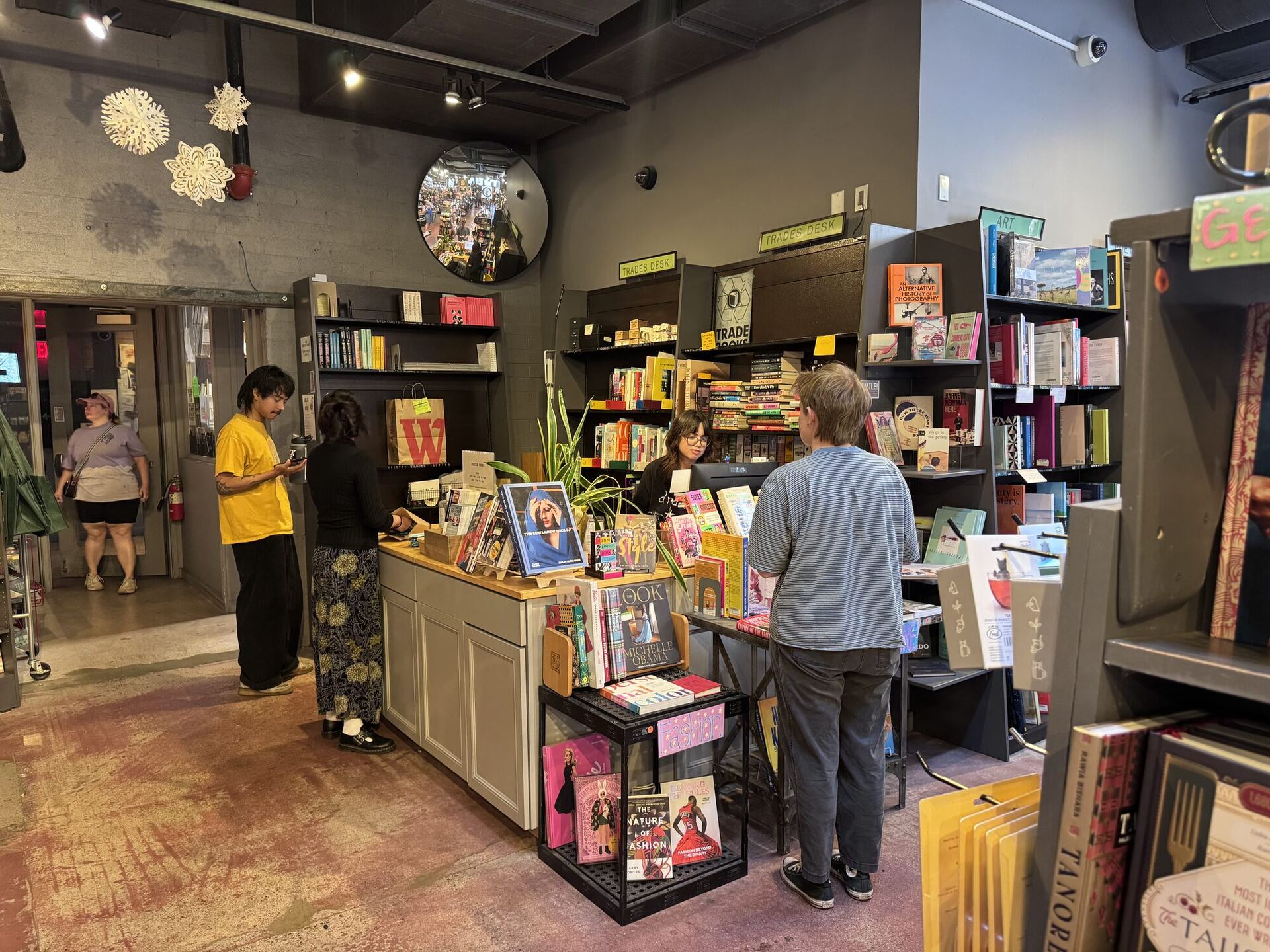 The trades desk, busy. A bookseller in a yellow T-shirt talks with two customers at the counter while another bookseller works behind it, under yellow TRADES DESK signs and a convex security mirror. Shelves of stationery and gifts crowd in on every side.