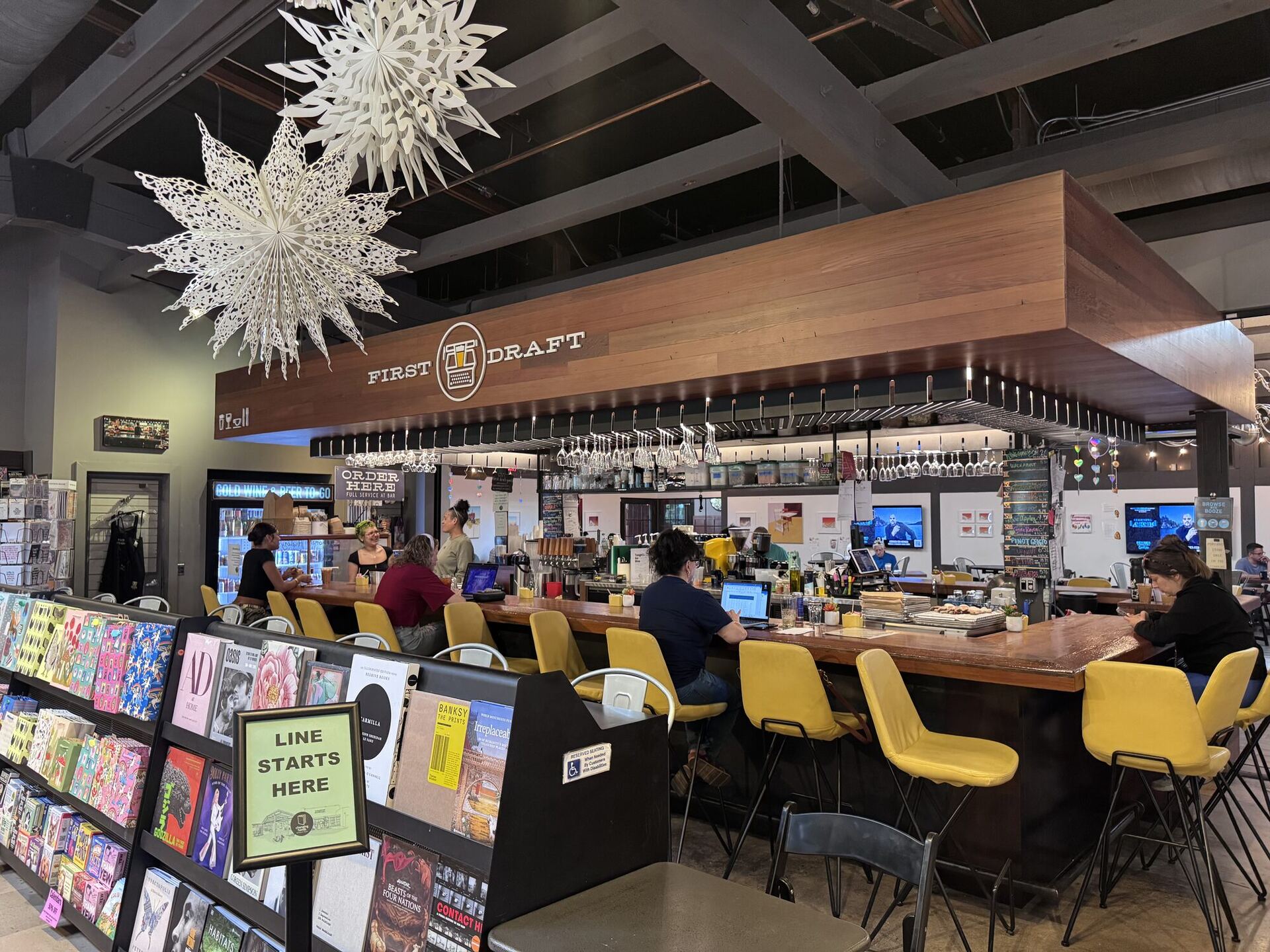 The FIRST DRAFT book bar: a long timber counter under a wooden canopy with wine glasses racked overhead, lined with yellow stools. Several people sit working on laptops, a barista pulls drinks, and a sign by the magazine racks reads LINE STARTS HERE. Two enormous paper snowflakes hang from the beams.