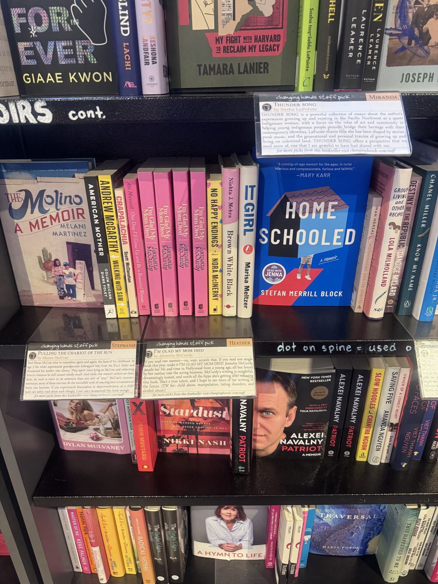 A memoir shelf where nearly every book carries a typed changing hands staff pick card signed by a bookseller. A handwritten strip of shelf tape reads MEMOIRS cont., and another reads dot on spine = used.