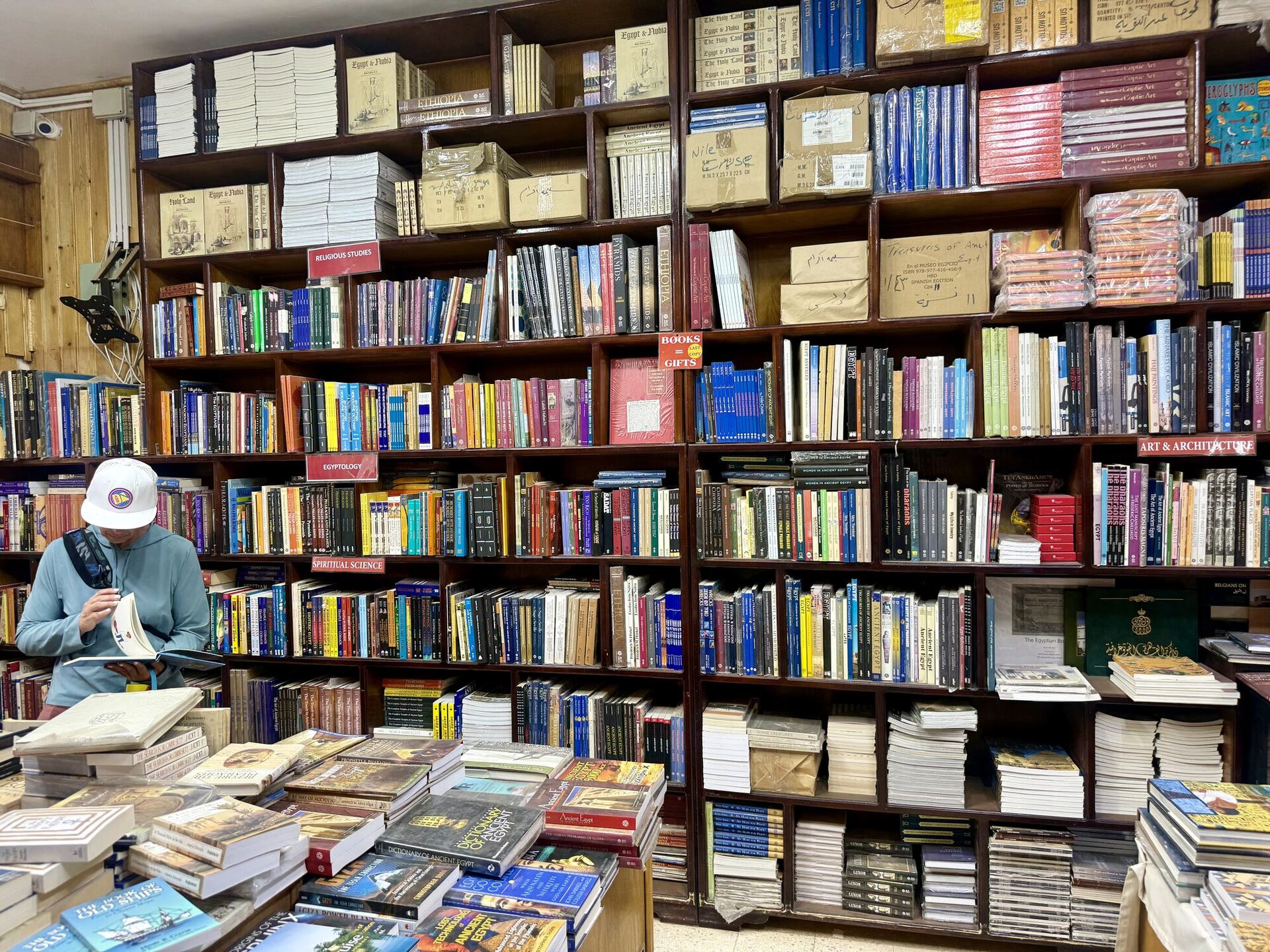 A wall of dark wooden pigeonholes from floor to ceiling, every compartment stacked and bundled with books and wrapped parcels. Small red cards mark the sections: EGYPTOLOGY, RELIGIOUS STUDIES, SPIRITUAL SCIENCE, ART & ARCHITECTURE. A customer in a white cap stands at the left reading, and tables of stock fill the foreground.