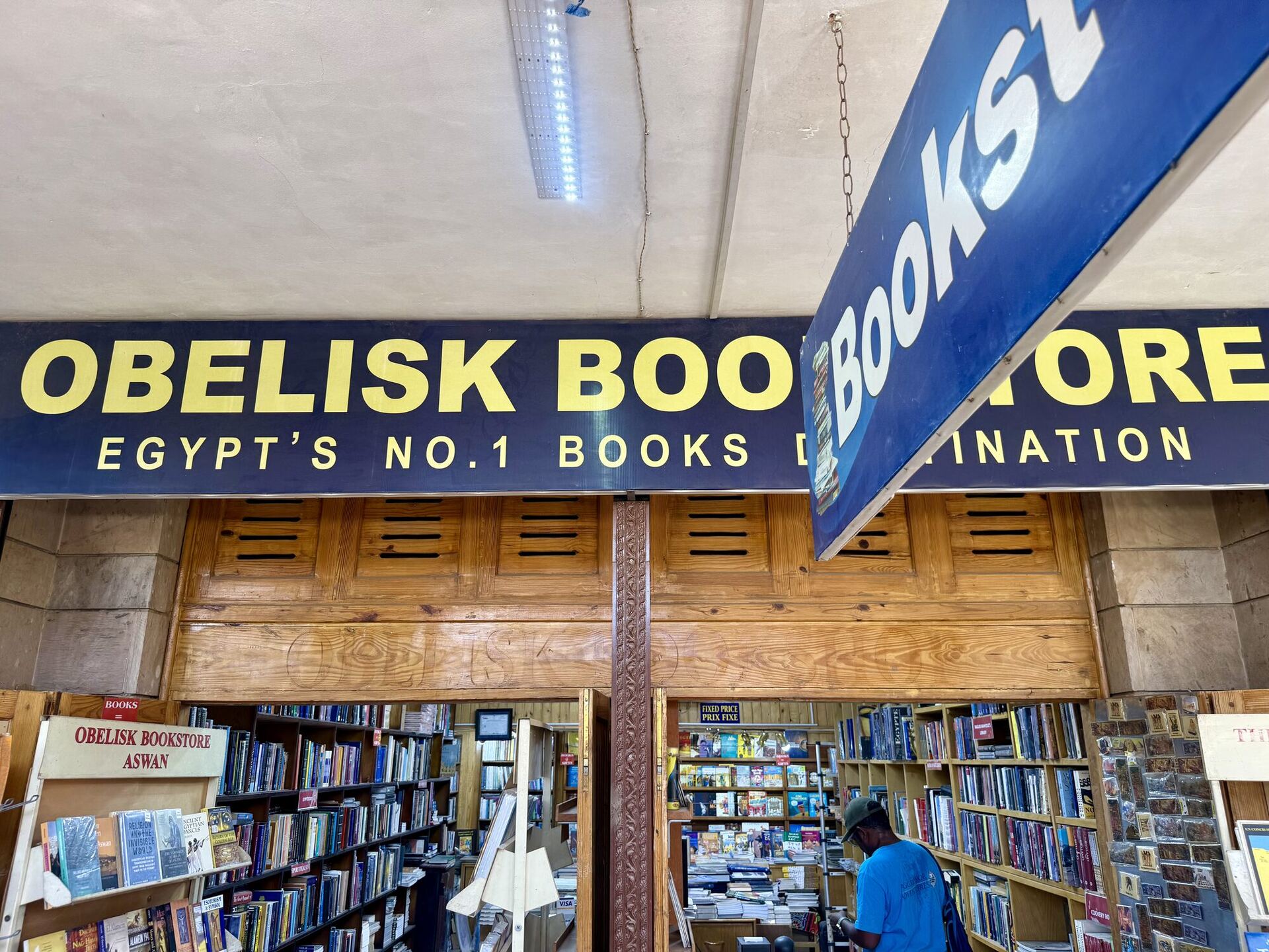 A long navy fascia board in yellow capitals reading OBELISK BOOKSTORE, EGYPT'S NO.1 BOOKS DESTINATION, with a second blue Bookstore sign swinging on a chain in front of it. Below, carved wooden shutters frame an open doorway into aisles of books, and a small painted board reads OBELISK BOOKSTORE ASWAN.