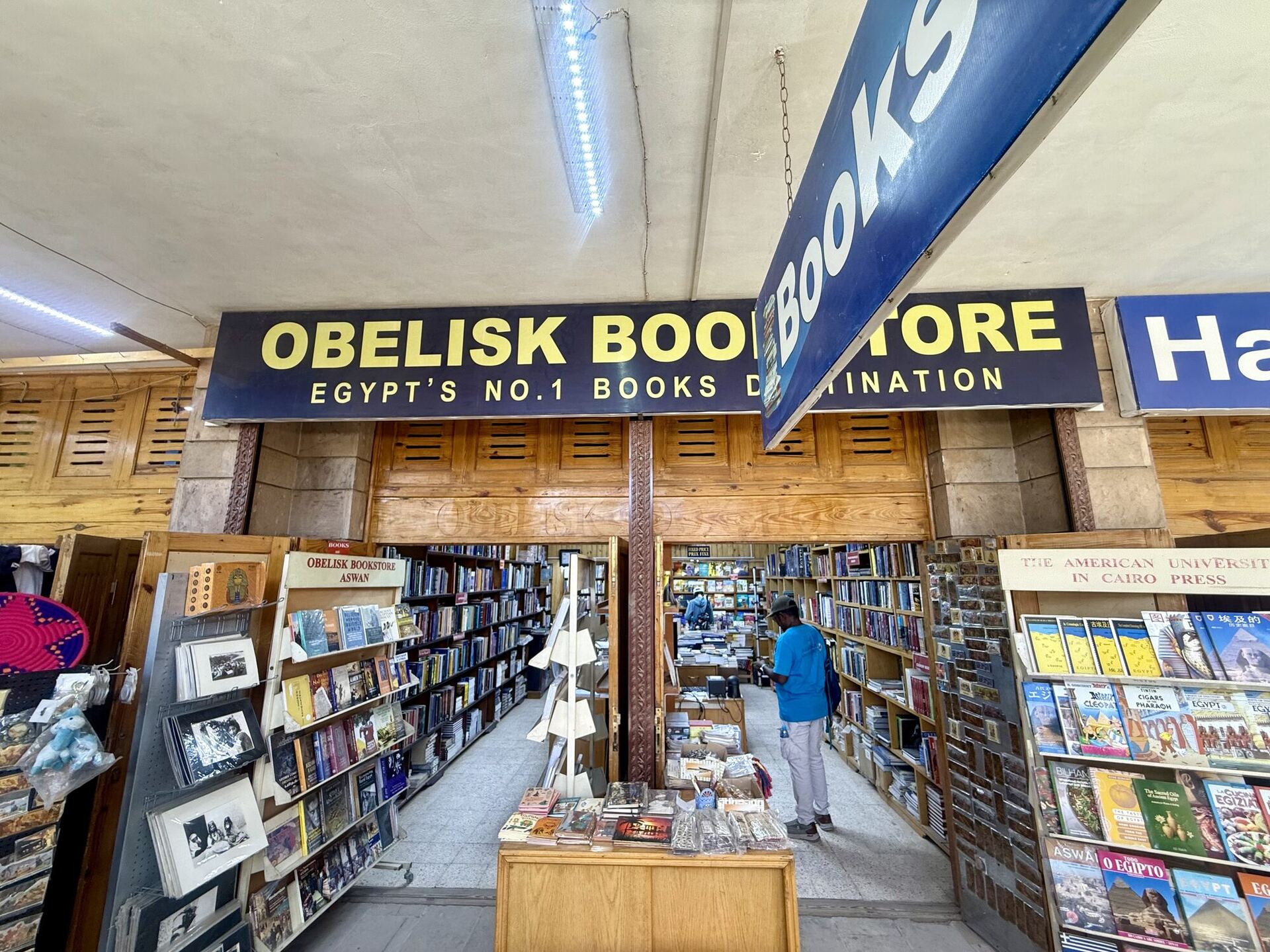 Looking out from inside the shop, under the same OBELISK BOOKSTORE fascia. Racks of postcards and guidebooks stand either side of a central aisle, a spinner of American University in Cairo Press titles fills the right of the frame, and a customer in a blue shirt browses at the back.