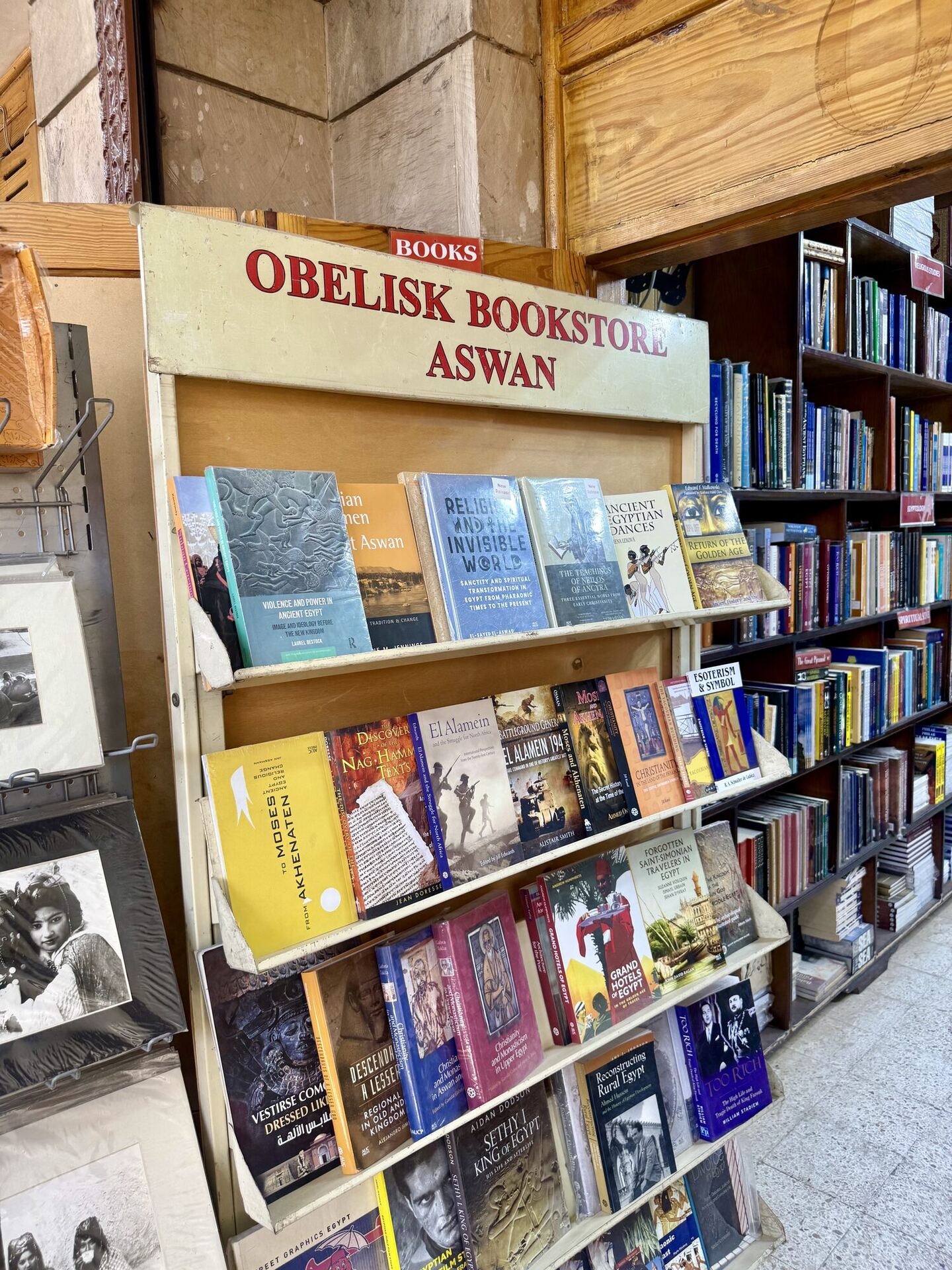 A cream-painted display stand headed OBELISK BOOKSTORE ASWAN in red capitals, tiered with books on Egyptology and Egyptian history: Religion and the Invisible World, Ancient Egyptian Romances, The Moses from Akhenaten, Rediscovering Rural Egypt.