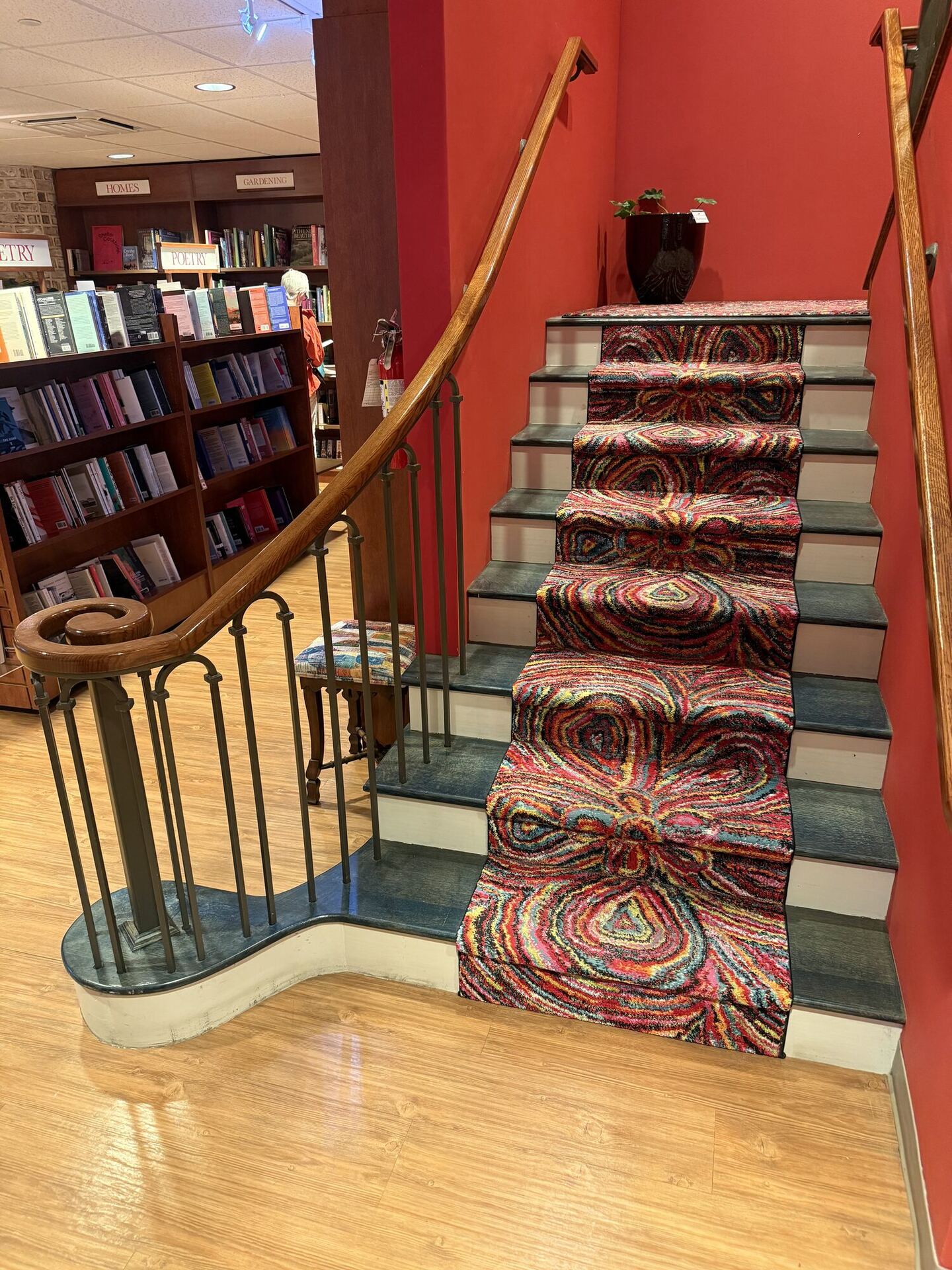 A staircase against a deep red wall, its treads covered by a runner of swirling multicoloured rag rug, with a curved wooden banister and slim iron balusters sweeping down to a scrolled newel post. Shelves under POETRY, HOMES and GARDENING signs stand at the foot of the stairs.