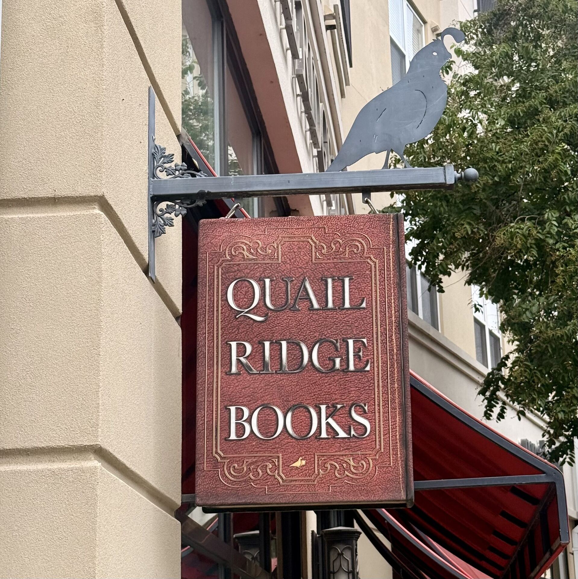 A projecting shop sign shaped like a red leather book, tooled with a gilt border and lettered QUAIL RIDGE BOOKS, hanging from an iron bracket with a cut-metal quail perched on top. A red awning and a street tree fill the background.