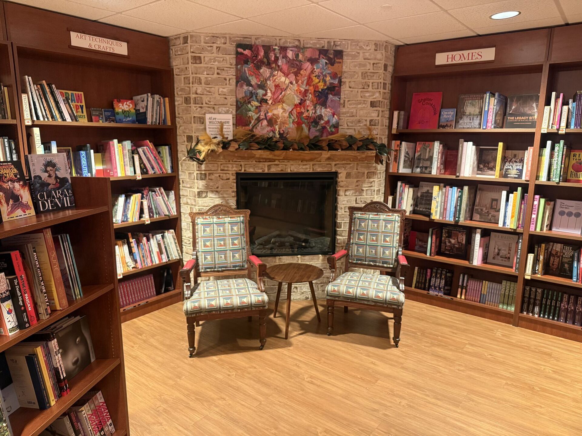 A fireplace nook. A pale brick chimney breast with a bright abstract painting over the mantel, two upholstered armchairs in a patchwork print drawn up to a small round table, and warm wooden shelving on both walls under section signs reading ART TECHNIQUES & CRAFTS and HOMES.