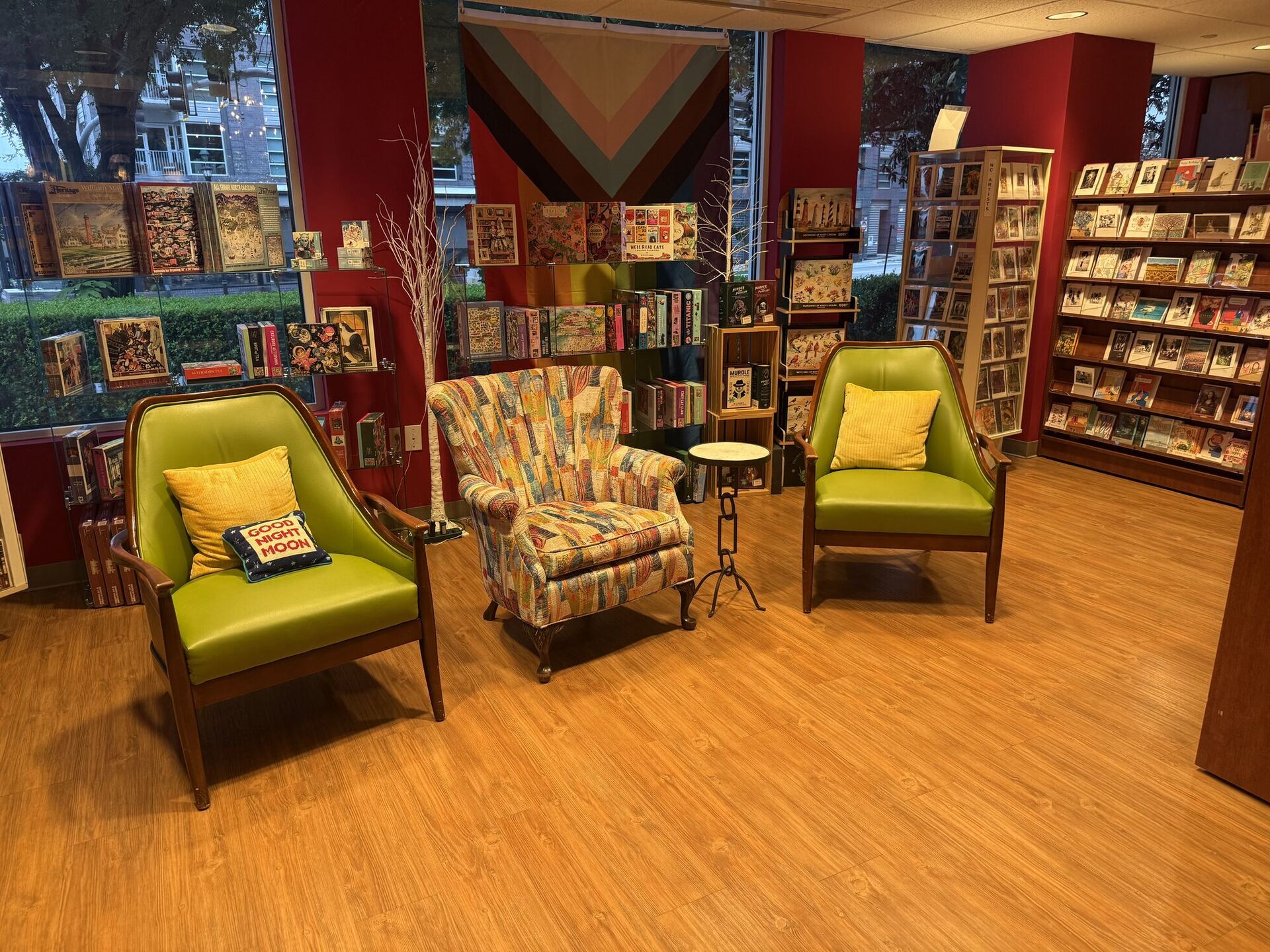 Three armchairs on a wooden floor in a window bay: two with lime-green vinyl upholstery and gold corduroy cushions, and between them a wing chair covered in a patchwork of old book covers. A Goodnight Moon cushion sits on the left-hand chair. Behind them, jigsaw puzzles and greeting cards fill the window ledges and a chevron banner hangs on the red wall.