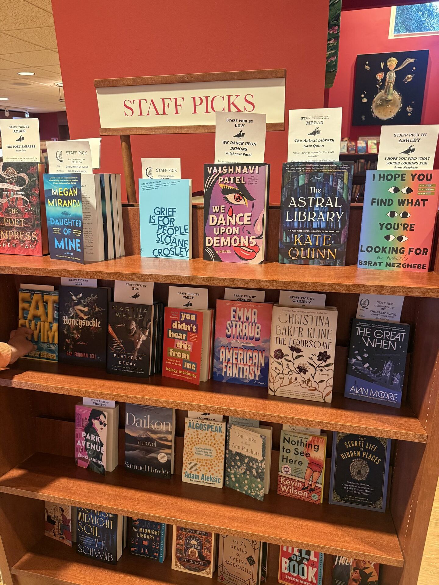 A tall wooden case under a hanging banner lettered STAFF PICKS in red. Face-out novels stand on every shelf, each with a printed card beside it headed STAFF PICK BY and a bookseller's first name, and a child's hand reaches into the frame at the left.