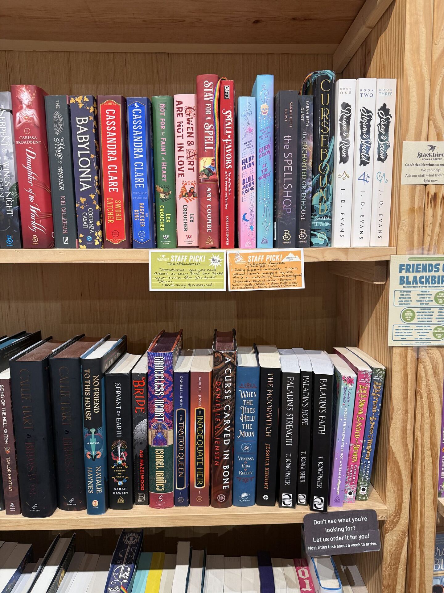 Two shelves of fantasy and romantasy on pale timber, dense with colour, with handwritten yellow STAFF PICK! cards tucked between them. A wooden sign at the right reads Blackbird Books & Coffee, Can't decide what to read? Ask our staff what they're reading right now, and a card below offers to order anything in about a week.