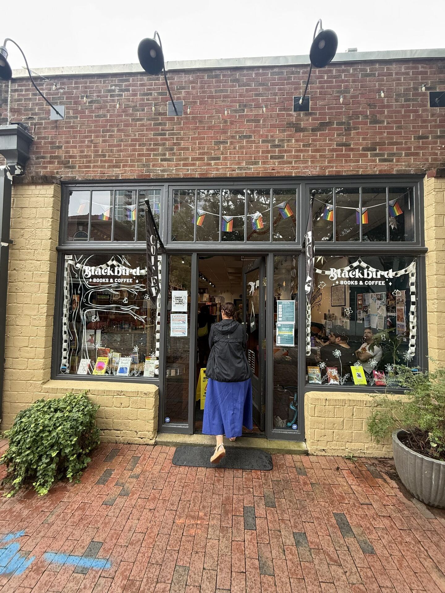 A brick and yellow-painted storefront with tall dark-framed windows, hand-lettered on the glass with a bare tree and the words Blackbird, Books & Coffee. Rainbow bunting hangs inside the transom windows, a COFFEE banner juts from the wall, and a customer in a black anorak and blue skirt steps through the open door.