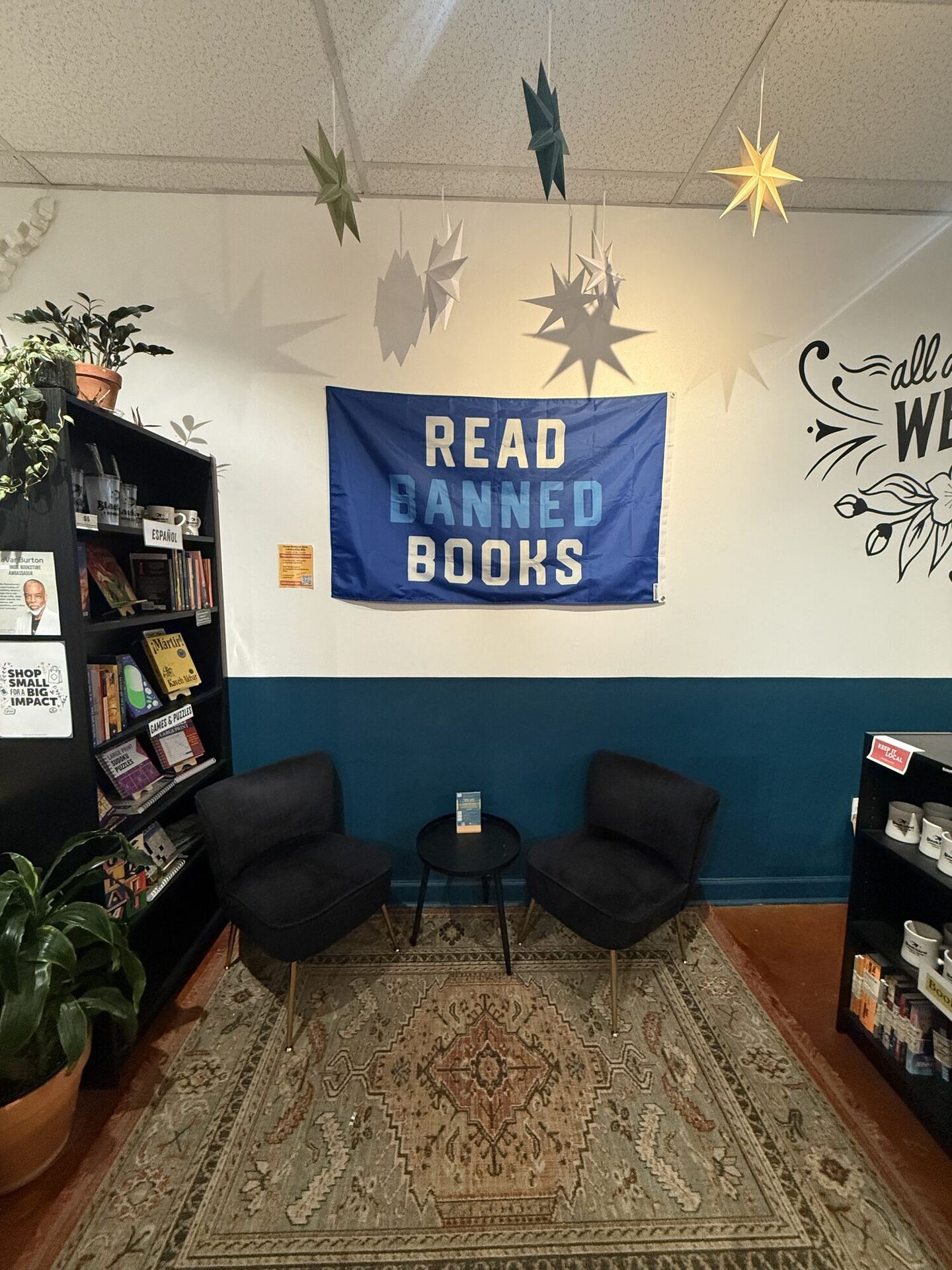 A corner with a cream wall above and teal below, hung with a blue flag reading READ BANNED BOOKS. Two black velvet chairs face each other across a small round table on a faded patterned rug, paper stars hang from the ceiling, and potted plants stand on a black bookcase to the left.