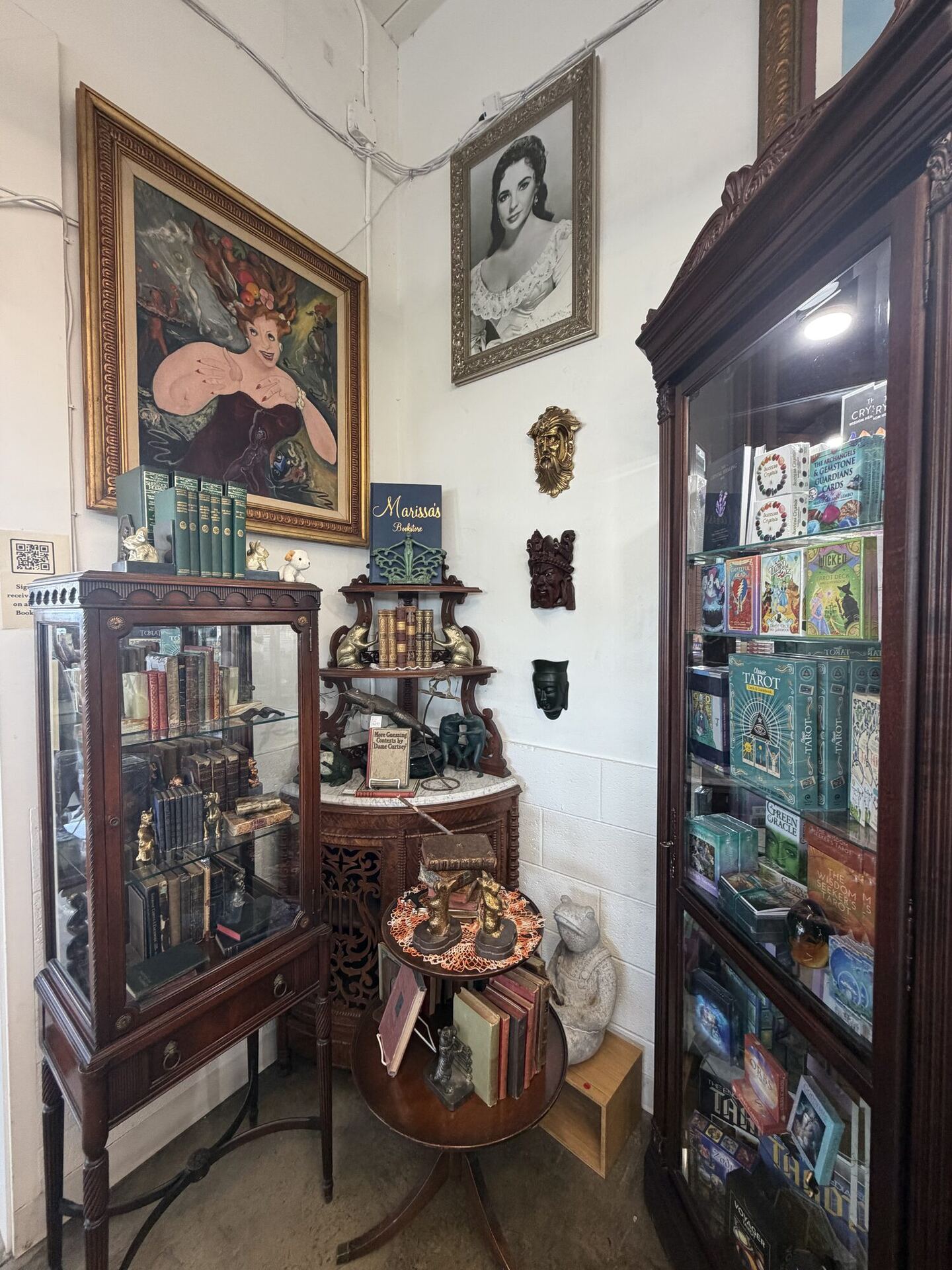 A crowded corner of curiosities: a gilt-framed burlesque oil painting and a portrait of a film star on the white brick walls, carved masks between them, a mirrored vitrine of antiquarian sets, a marble-topped console stacked with old books, and a tall glass cabinet of tarot decks. A small navy sign on the shelf reads Marissa's Bookstore.