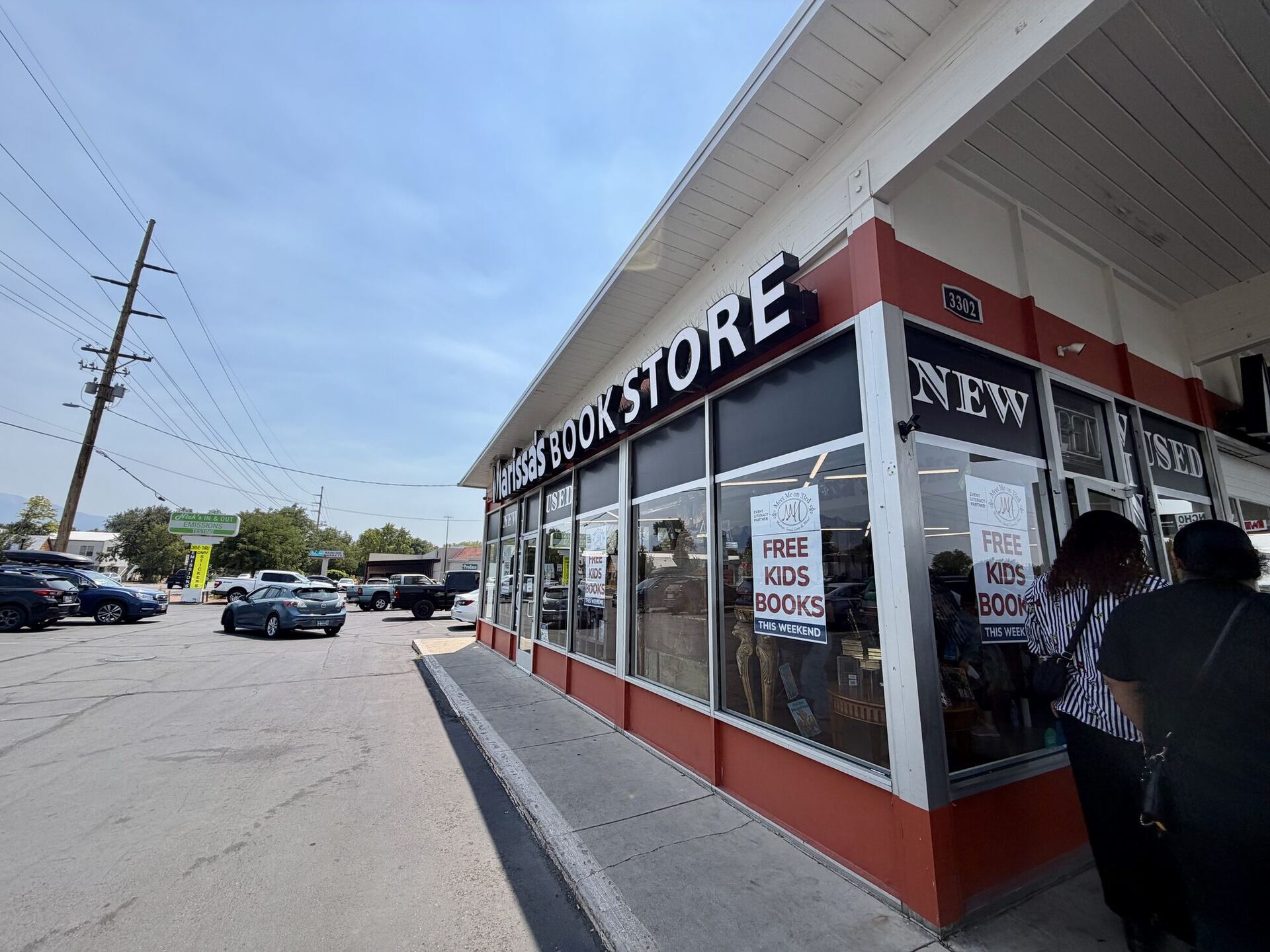 A low strip-mall corner unit under a white canopy, its rust-red base wall running back along a car park. Black channel letters on the fascia read Marissa's BOOKSTORE, the windows are lettered NEW and USED, and two hand-made signs promise FREE KIDS BOOKS THIS WEEKEND.