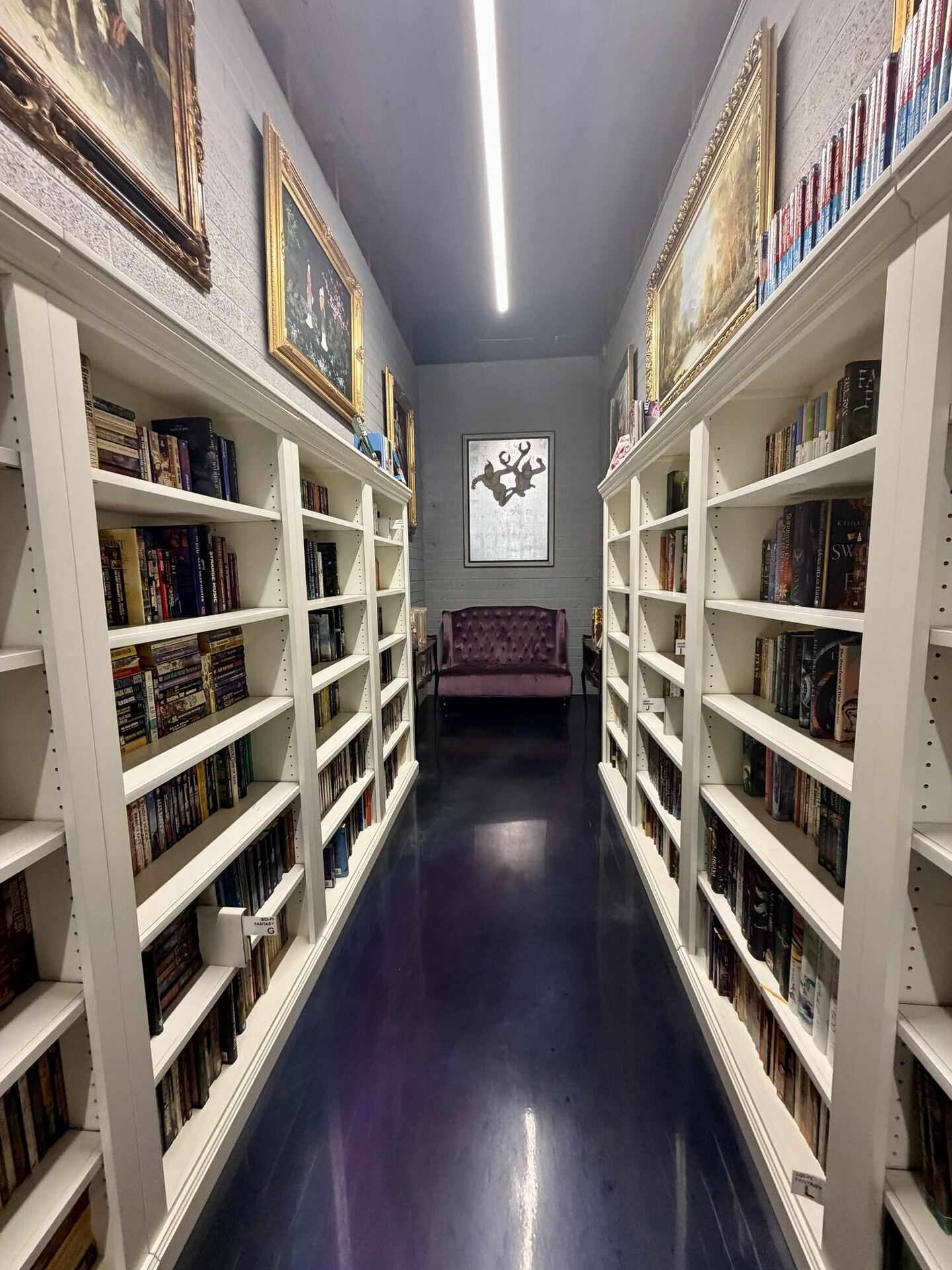 A narrow aisle between tall white bookcases packed to the edges, gilt-framed oil paintings propped along the tops of both runs, a strip light overhead, and a glossy deep purple floor leading to a plum velvet settee under a framed print at the far end.