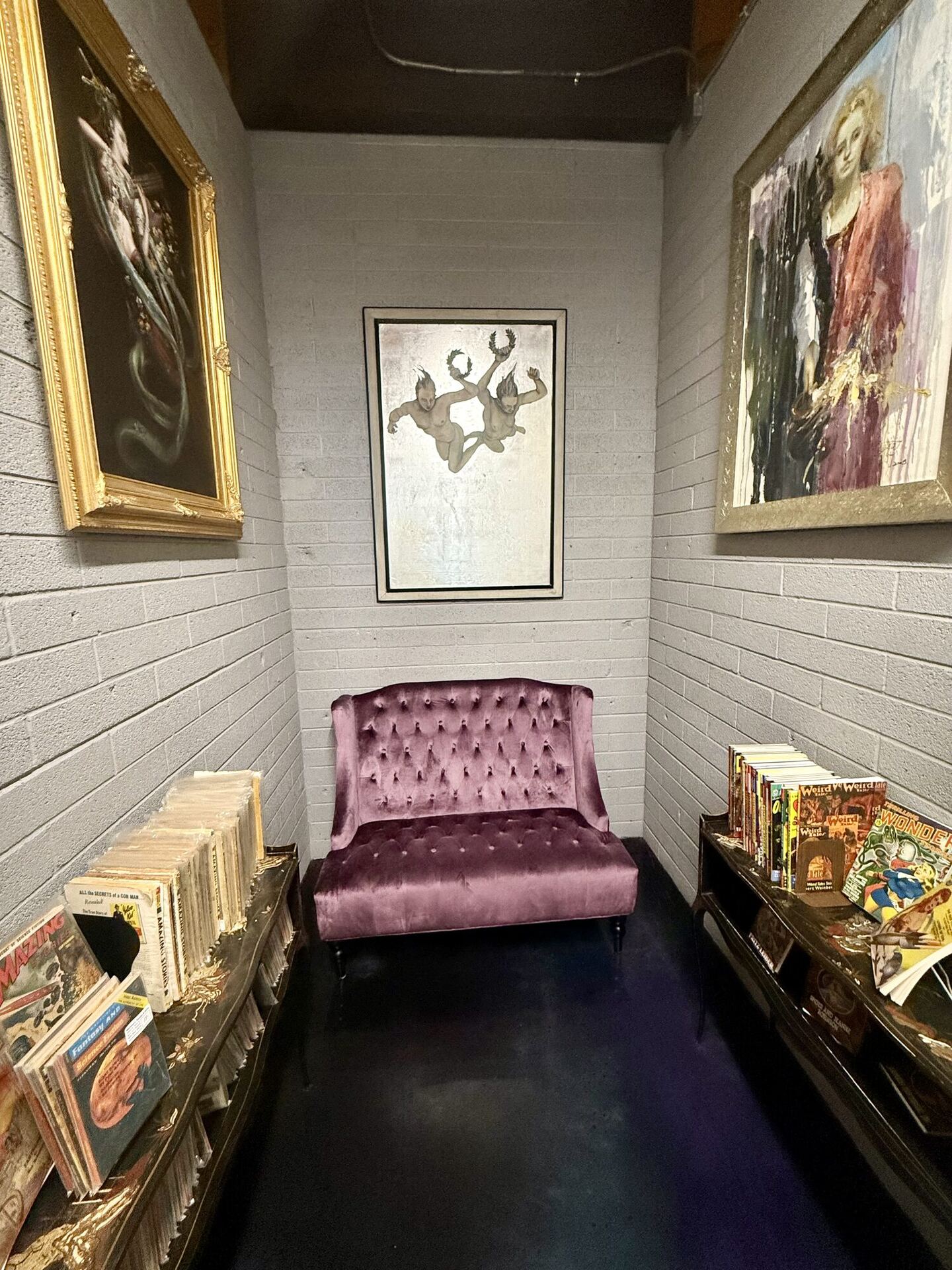 A white-painted brick alcove with a plum velvet button-backed settee against the end wall, a framed print of two tumbling figures above it, a large gilt-framed oil painting on each side wall, and long boxes of back-issue comics on stands to left and right.