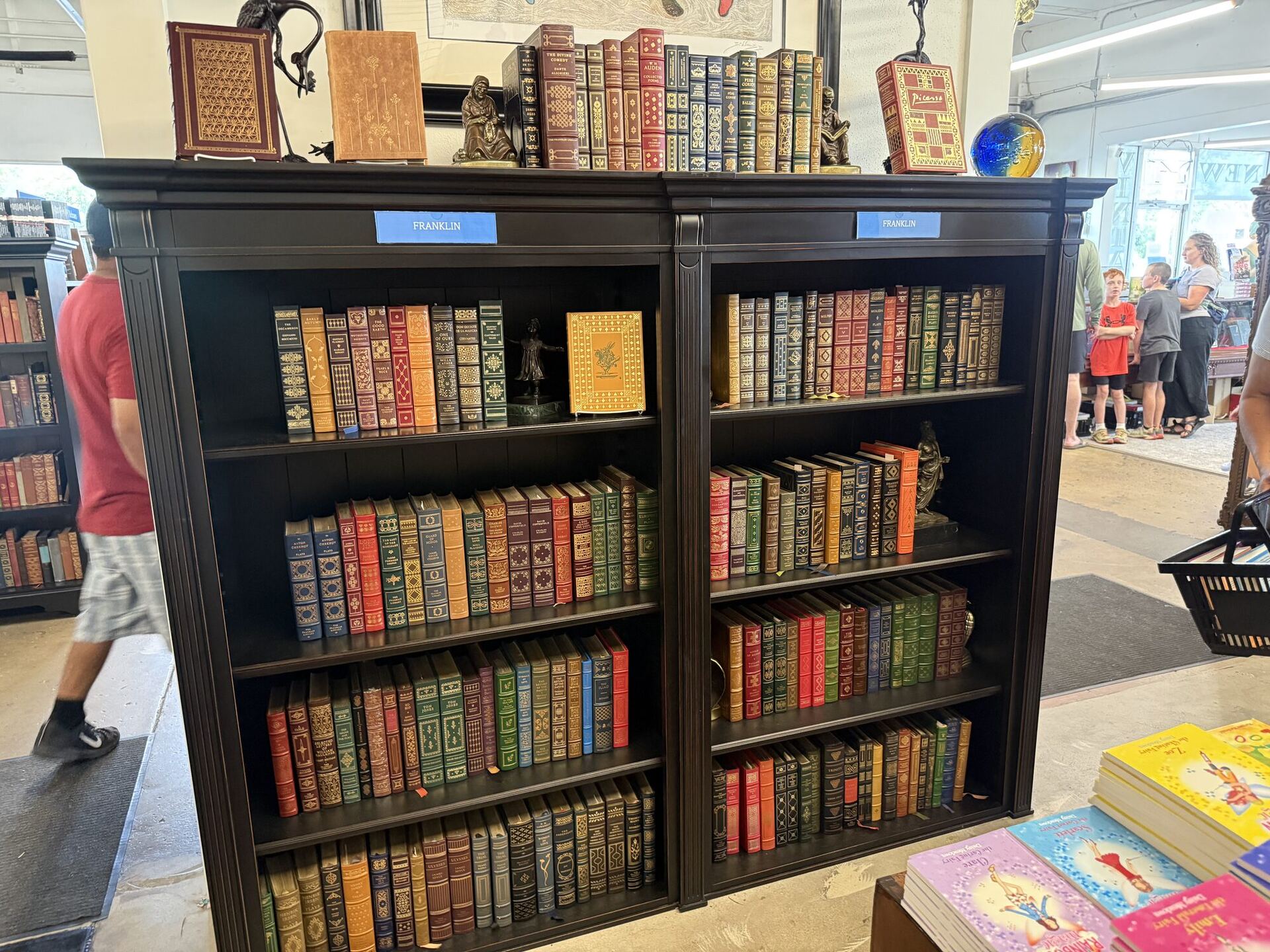Two matched black bookcases labelled FRANKLIN, filled with gilt-tooled Franklin Library leather editions in red, green and tan, with small bronze figures and a glass globe on top. Shoppers move through the aisles behind them.