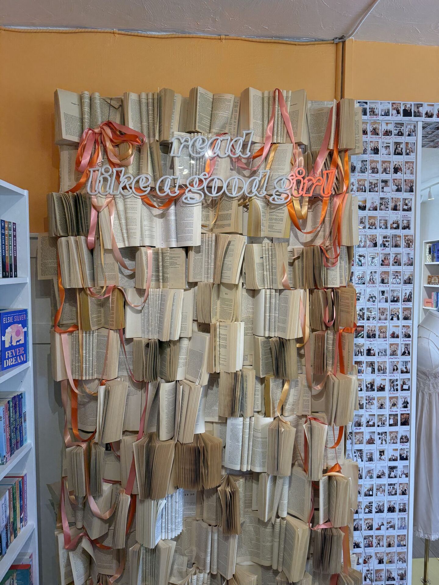A whole wall built from open paperbacks fixed page-out in overlapping rows, threaded with trailing pink and orange ribbons, with cut-out lettering across the middle reading read like a good girl. A grid of hundreds of instant photographs of customers covers the wall to the right.