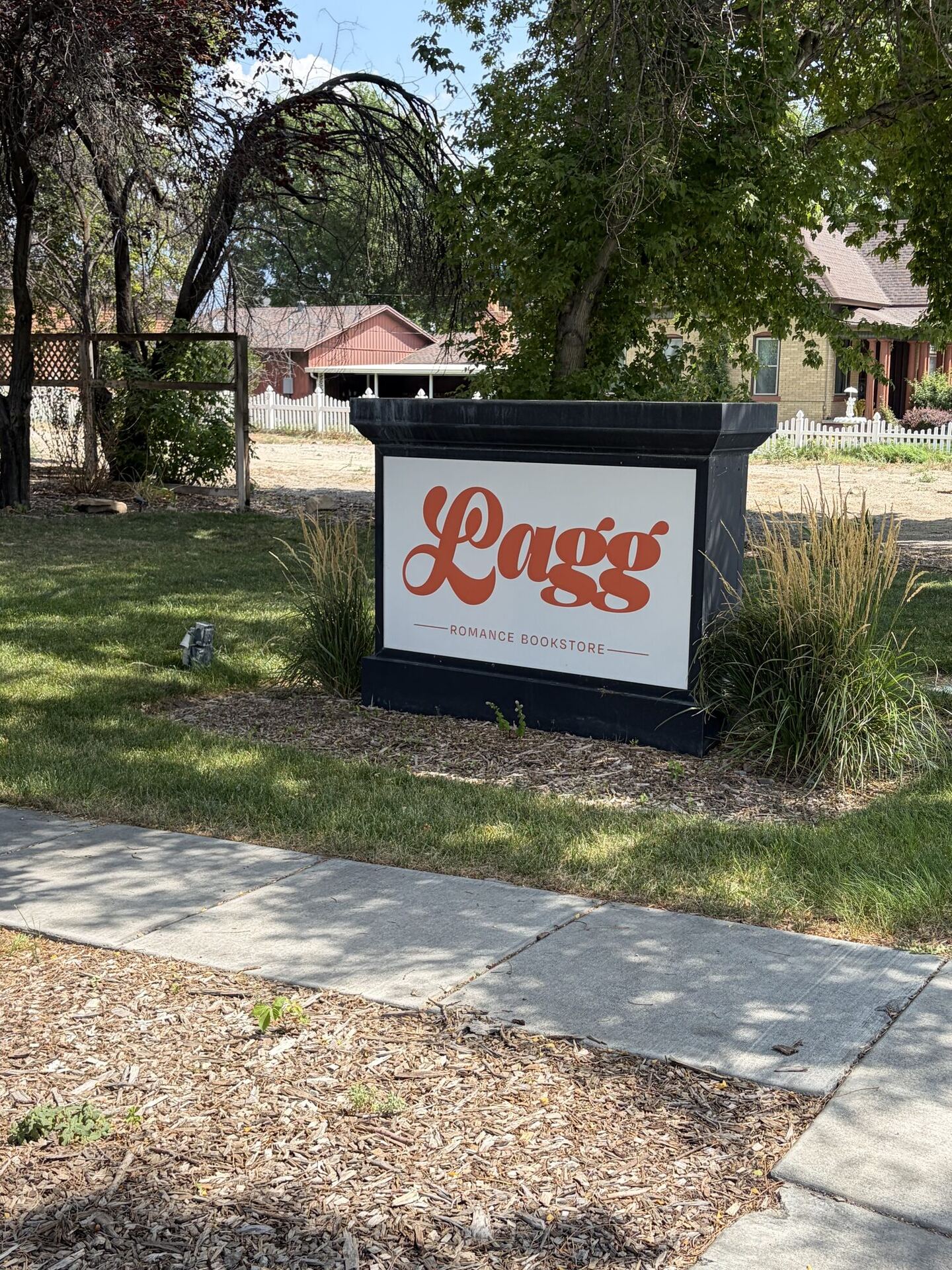 A low monument sign on a lawn under mature trees, its white face carrying Lagg in a curling burnt-orange script above the words ROMANCE BOOKSTORE, with ornamental grasses at its base and a white picket fence and houses behind.