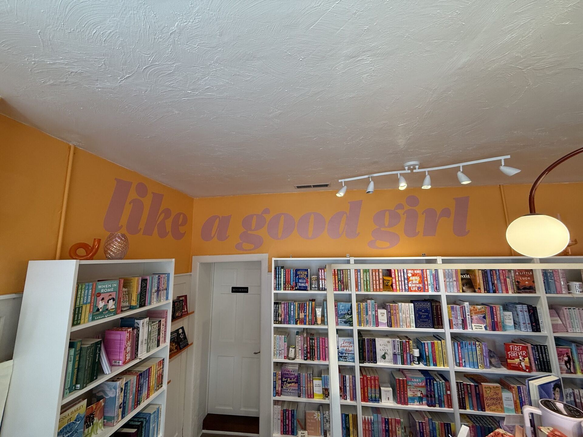 A room painted marigold above white panelling, with like a good girl painted large across the wall in lilac script. White shelves below hold rows of romance paperbacks, face-out and spine-out, lit by a track of spotlights and a globe lamp.