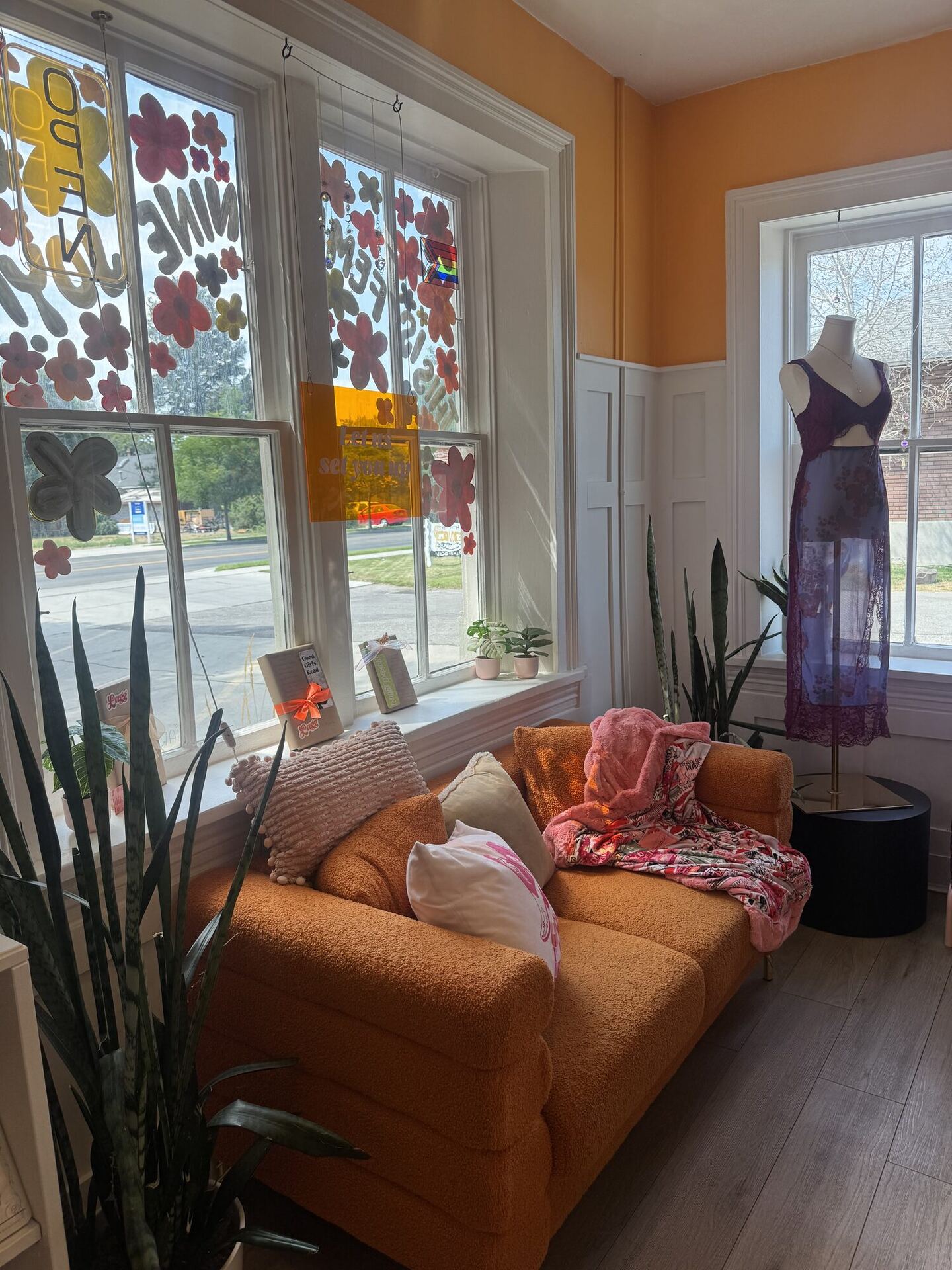 A burnt-orange bouclé sofa in a sunny window bay of an old house, heaped with pink and cream cushions and a floral throw. Painted flowers cover the sash windows, snake plants stand on the floor at either side, and a dressmaker's mannequin wears a plum lace slip dress beside a black side table.