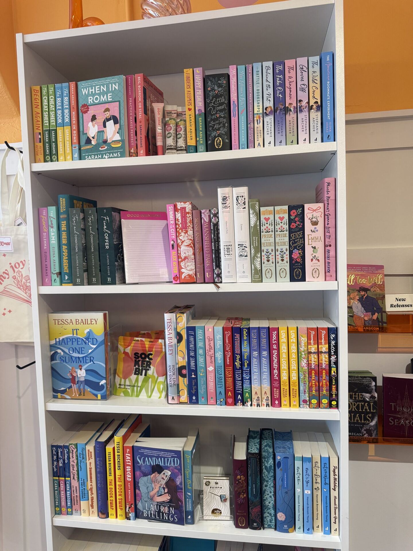 A white bookcase of romance against a marigold wall: Sarah Adams and Tessa Bailey shelved face-out, a shelf of pastel special editions and Austen reissues, notecards and a book-shaped tote hung at the side, and a New Releases card at the right.